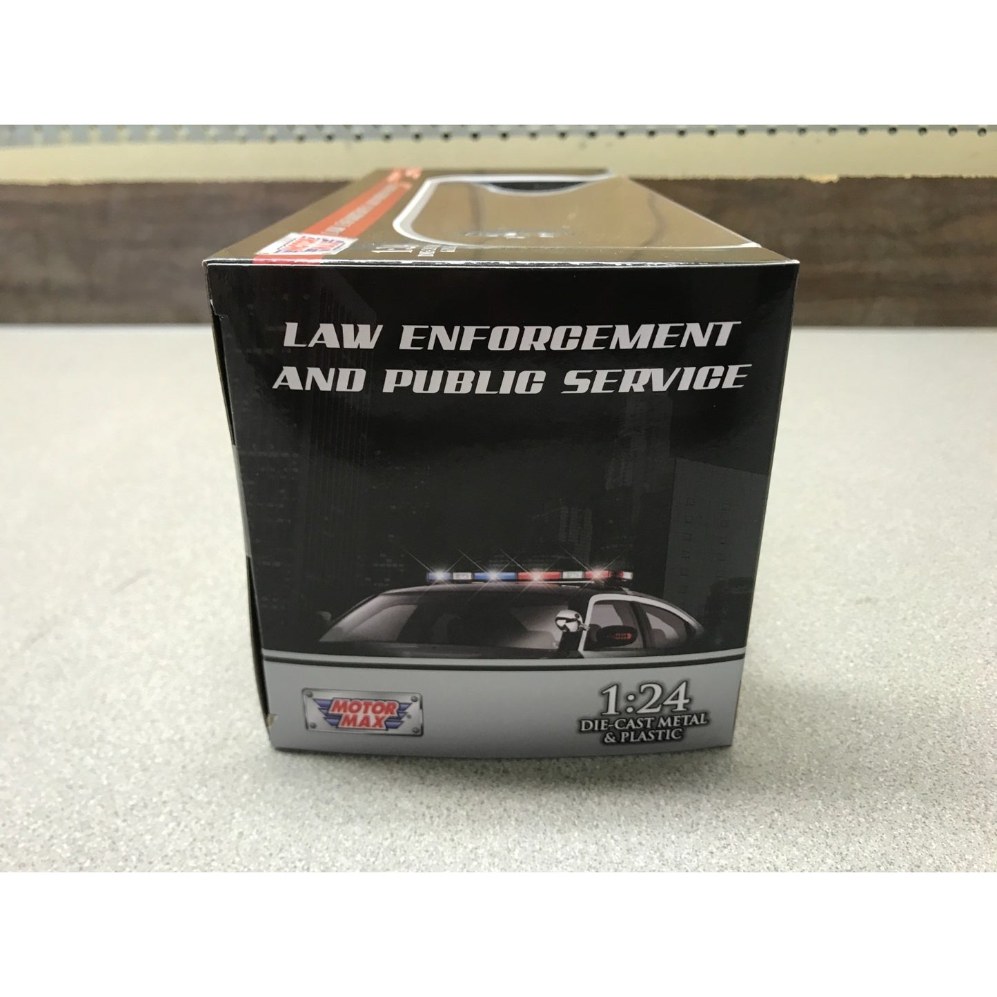 A boxed miniature model car on a light gray surface. The box is black with white text reading 'LAW ENFORCEMENT AND PUBLIC SERVICE' and shows a small police car with flashing lights.