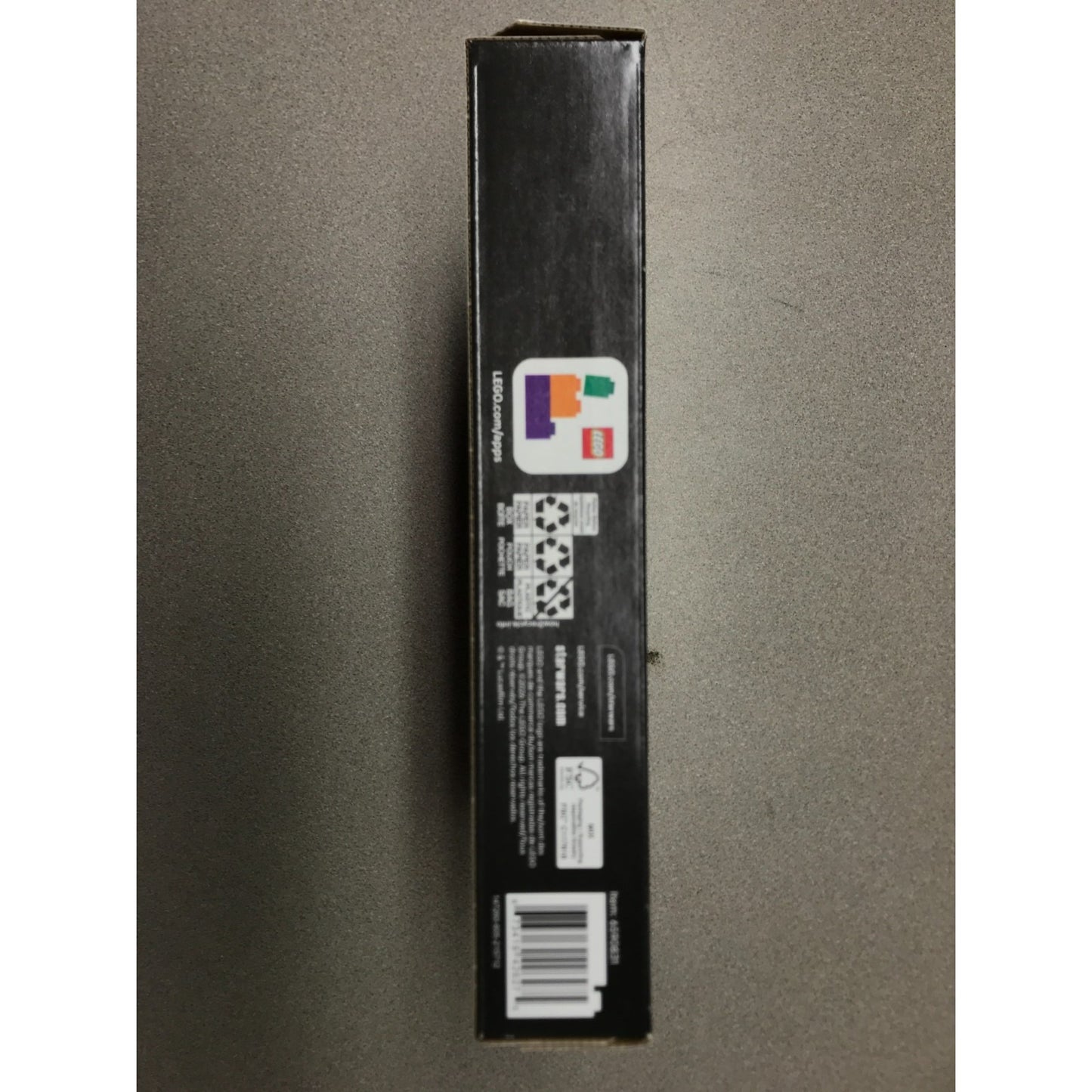 A tall, narrow black product box standing upright against a gray background, showing printed labels, a barcode, and a small multicolor logo near the top.