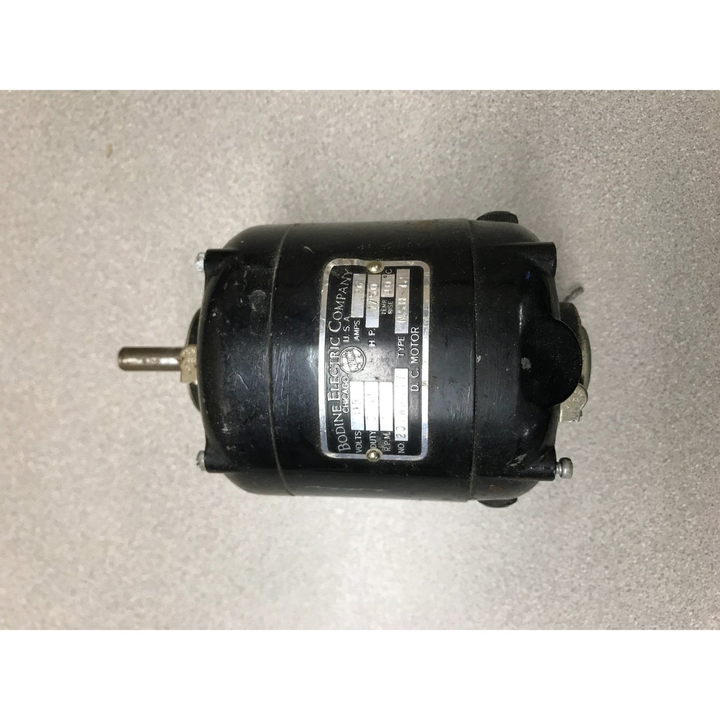 A vintage black electric motor with a metal nameplate, photographed on a light gray textured surface.