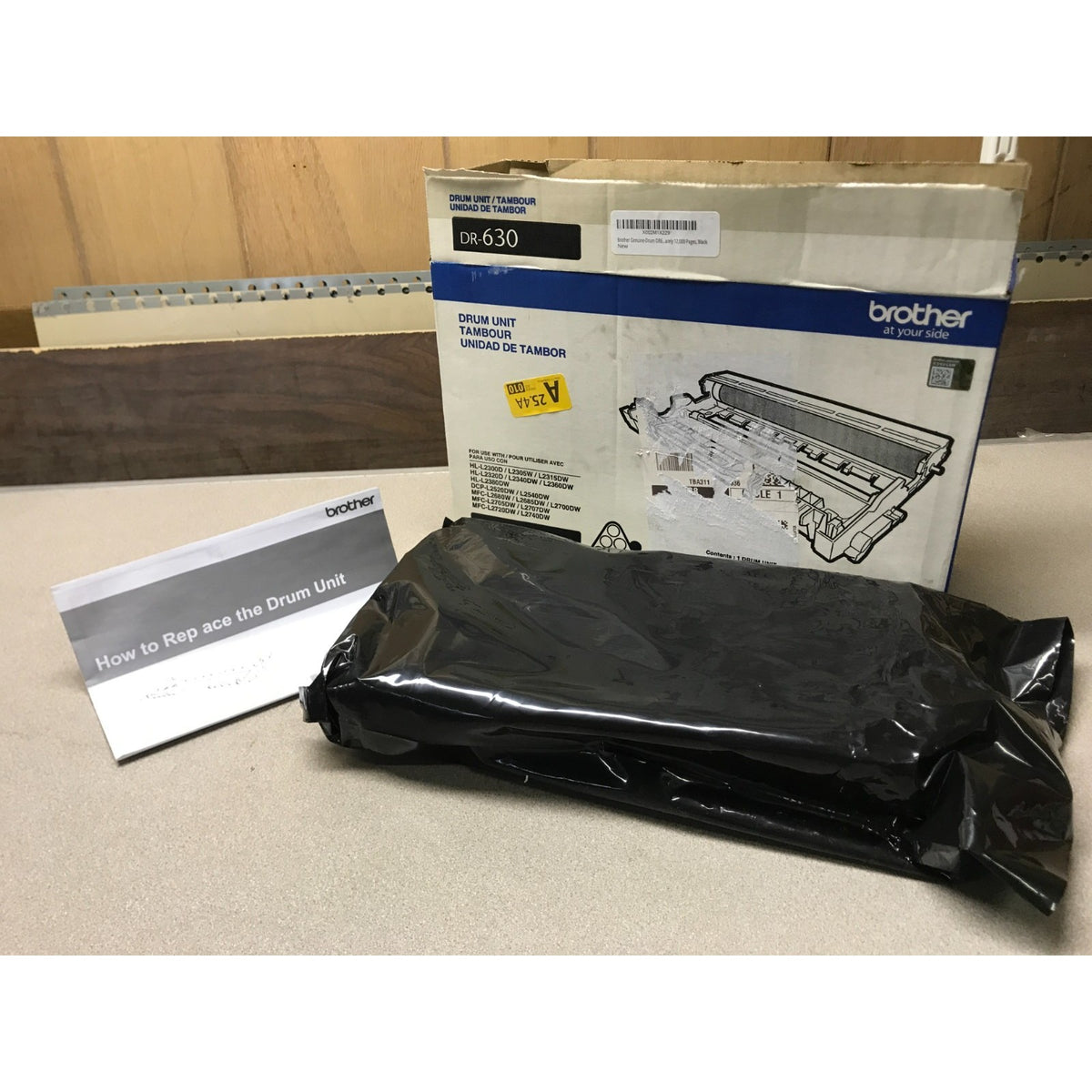 A Brother drum unit replacement kit including a black plastic-wrapped drum unit, its original retail box, and a small instruction card, set on a beige tabletop.