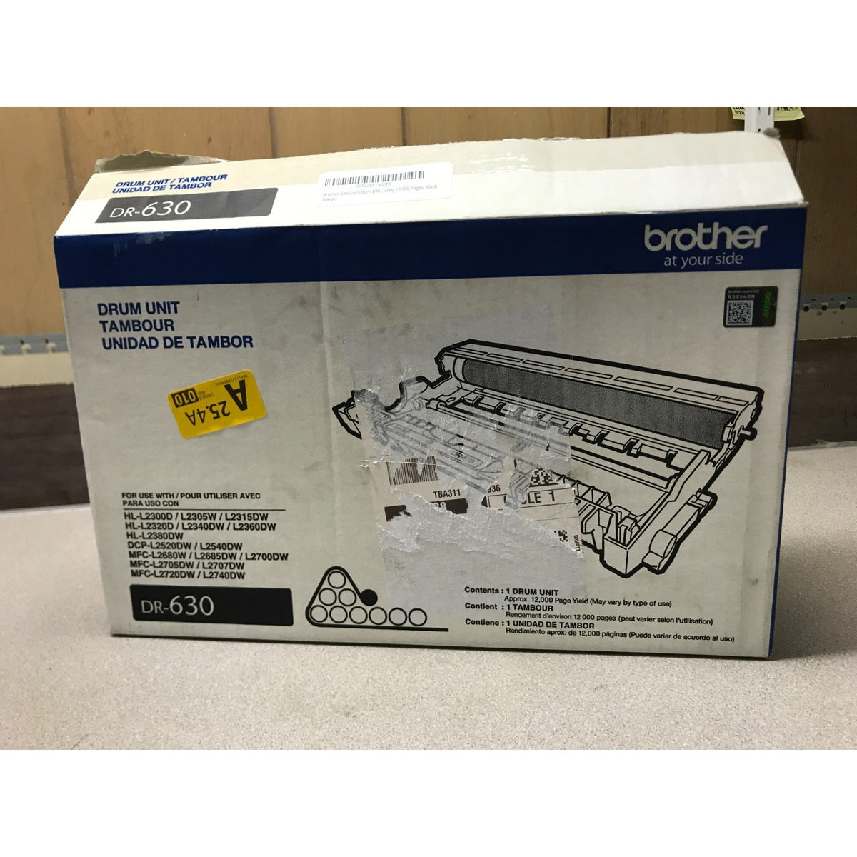 Brother DR-630 drum unit retail box shown on a light countertop with a wooden background. The white and blue packaging features product text and a printed illustration of the drum unit.