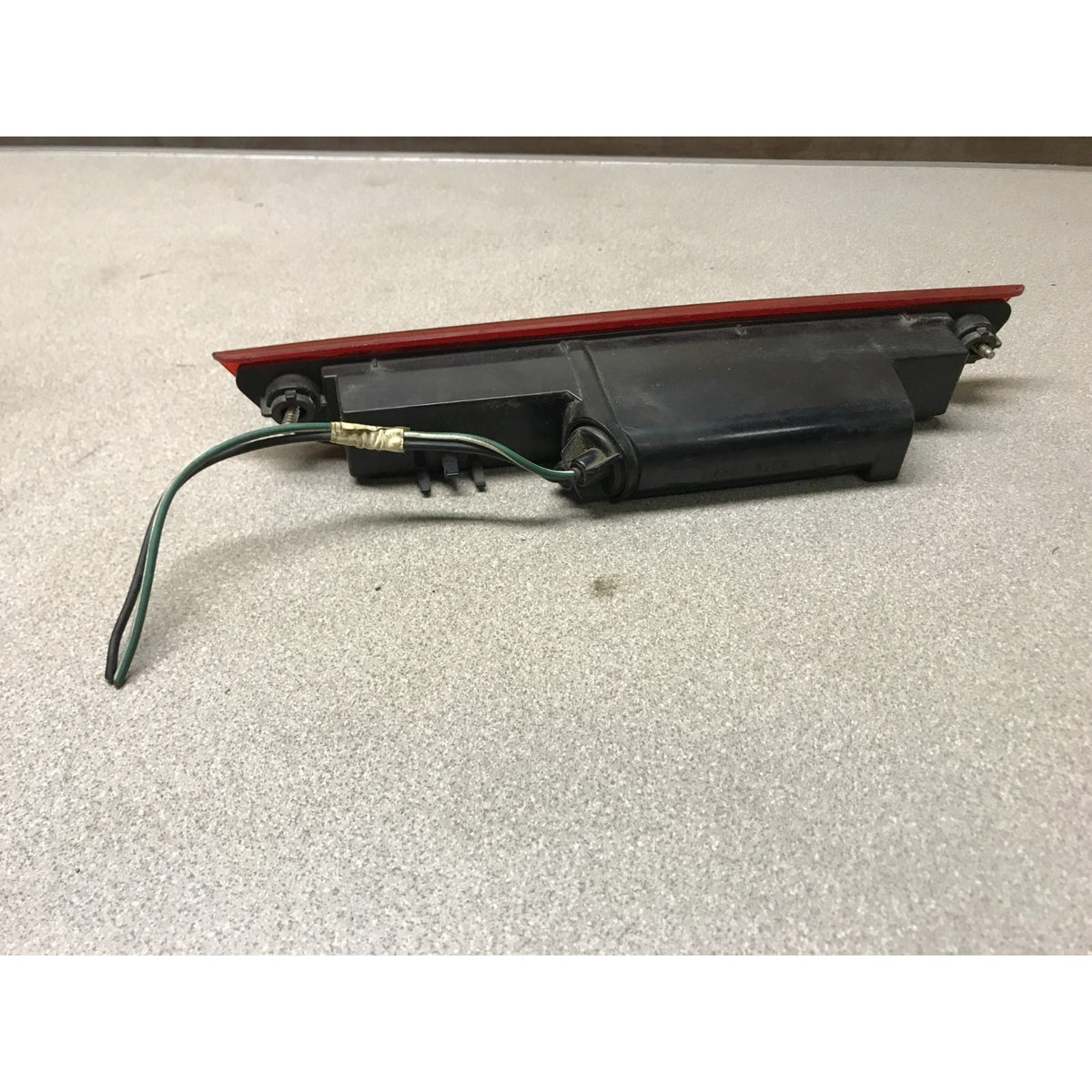 A used automotive light assembly, brake light, featuring a black plastic housing, a red reflective strip, and a green wire harness, resting on a gray textured floor. 2005-2010 Honda Odyssey OEM High Mount Third Brake Light back view