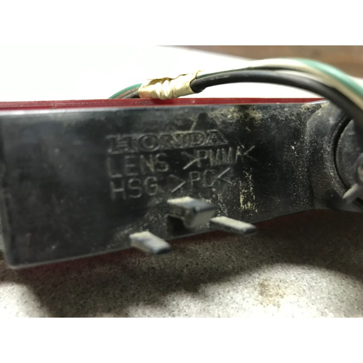 A close-up of a used black Honda lens assembly with visible pins, a gold-colored terminal wrap, and attached wires on a concrete surface. 2005-2010 Honda Odyssey OEM High Mount Third Brake Light