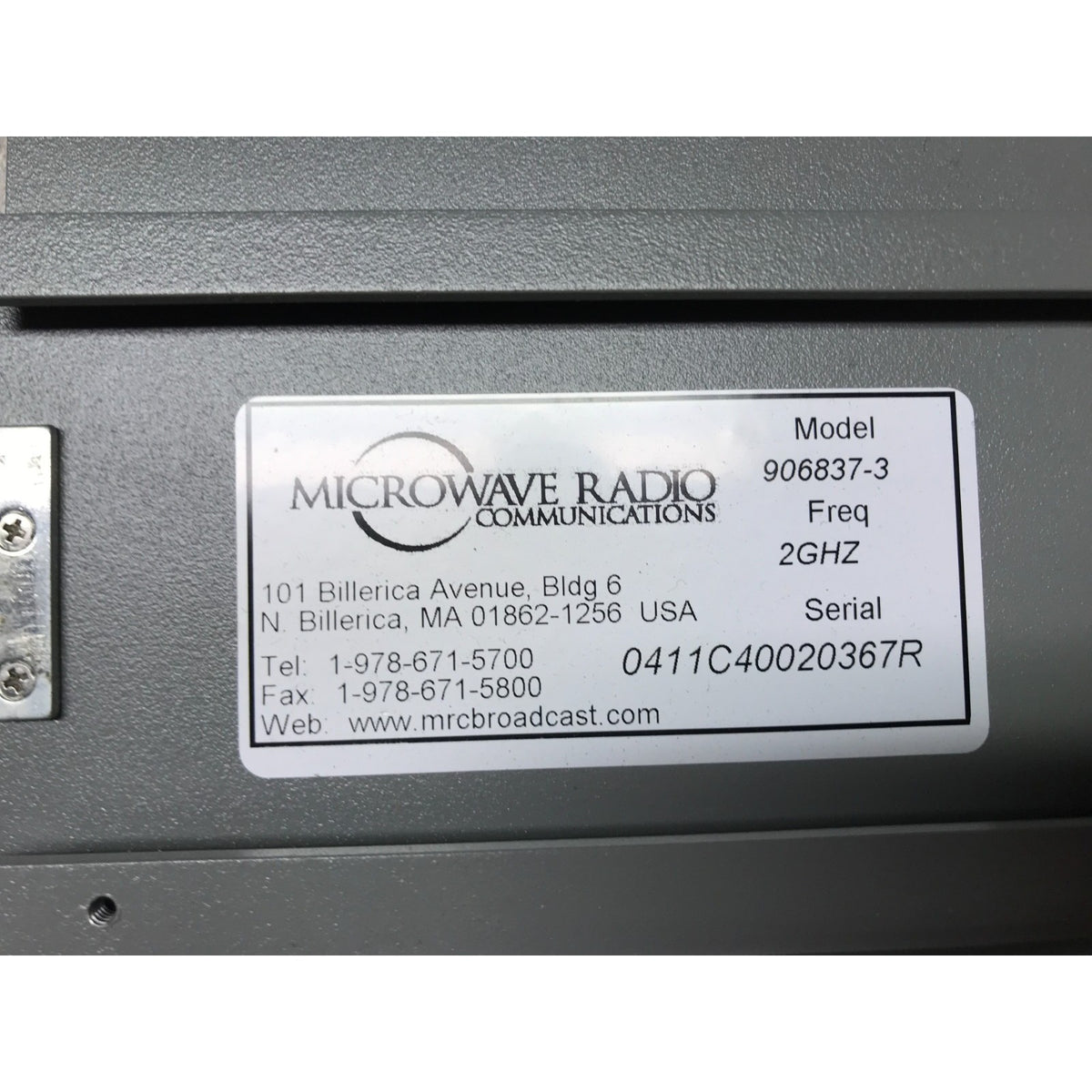 A close-up of a metal product label on a dark gray device. The label reads MICROWAVE RADIO COMMUNICATIONS and lists model 906837-3, freq 2GHZ, plus serial and contact details.