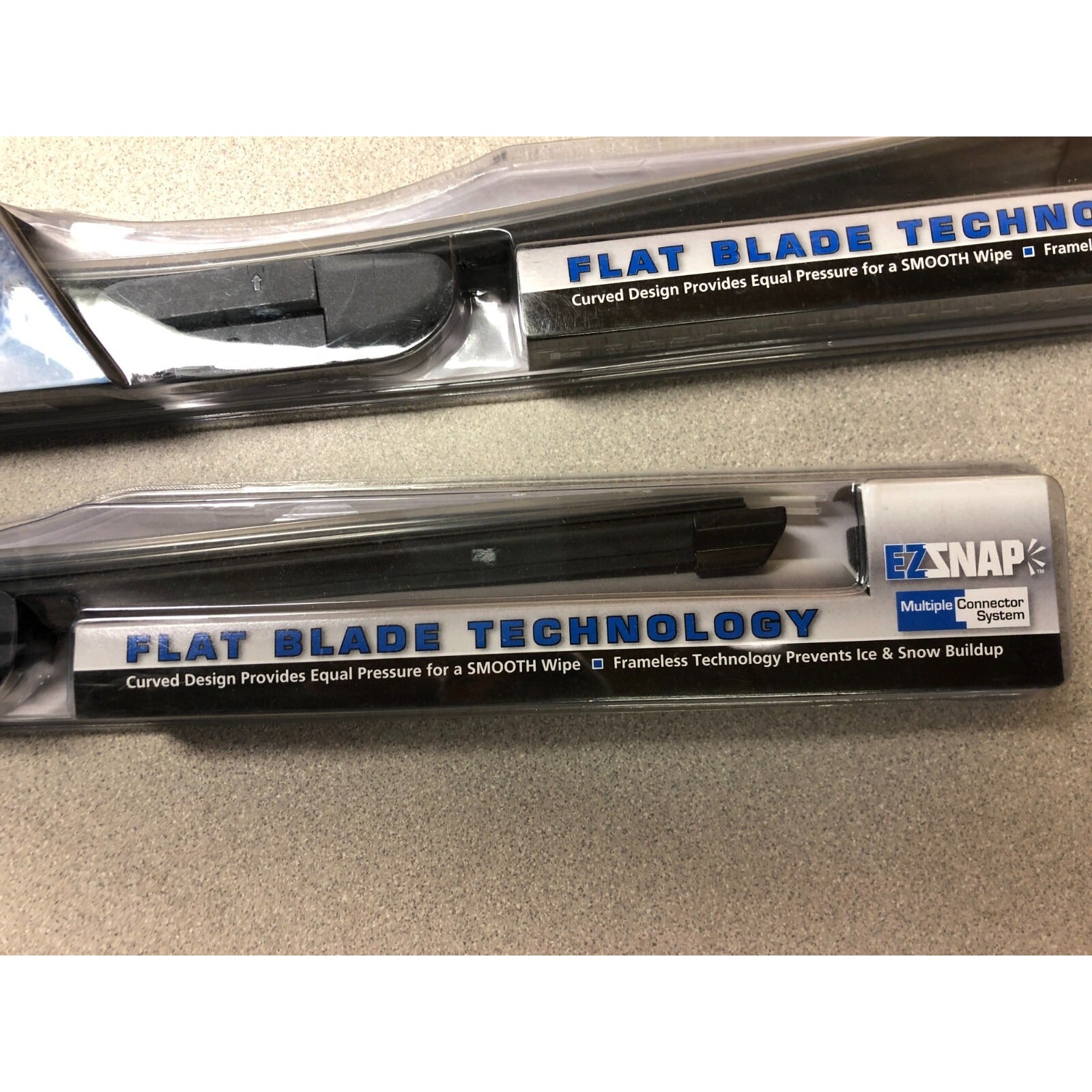 Two windshield wiper blades with 'Flat Blade Technology' labels on a beige surface. EXSNAP