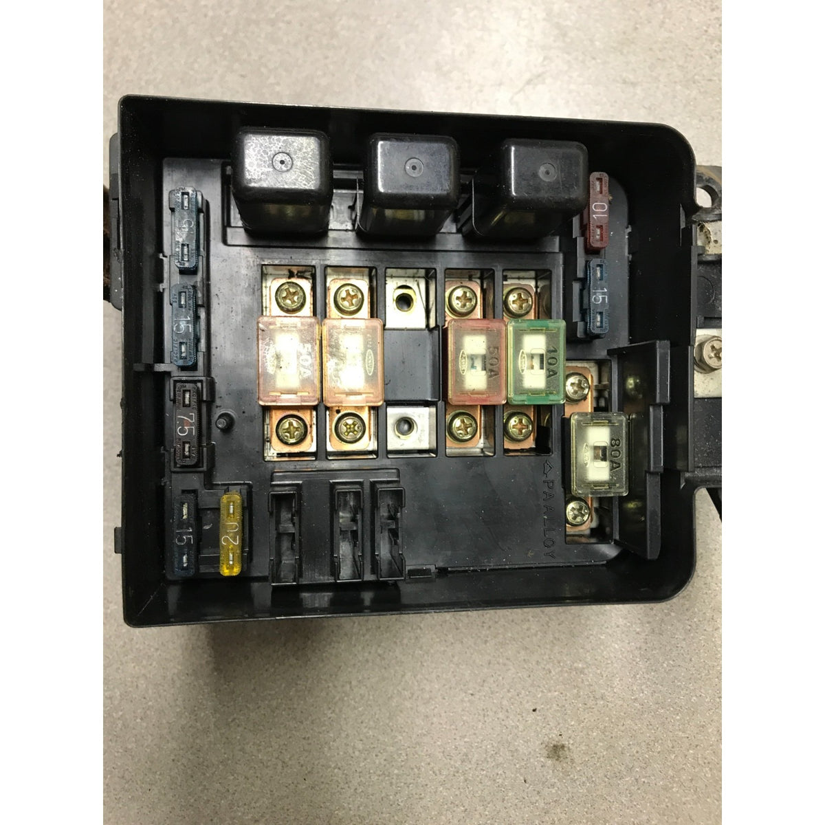 A black plastic automotive fuse box with multiple fuses and relays, photographed from above on a light gray floor. 1993 Honda Civic Del Sol OEM Engine Bay Fuse Box