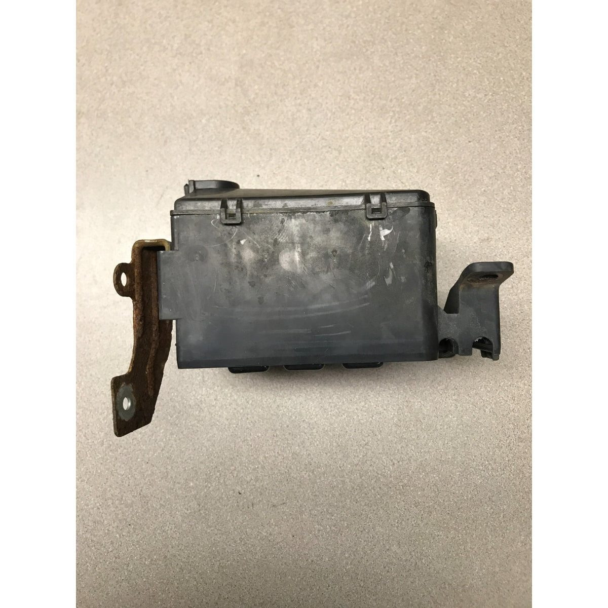 A used black plastic automotive component with a rusted brown metal bracket on the left side, photographed on a neutral beige textured surface. 1993 Honda Civic Del Sol OEM Engine Bay Fuse Box side view
