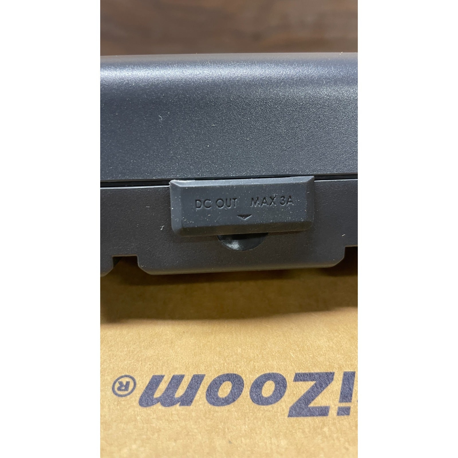 Close-up of a black electronic device with 'DC OUT MAX 3A' label on a brown surface with 'ZOOM' branding.