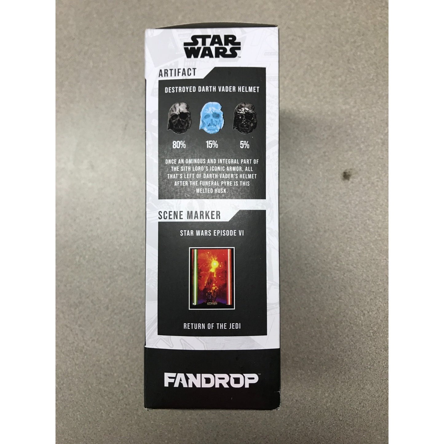 A Star Wars Fandrop collectible box for an 'Artifact' depicting a destroyed Darth Vader helmet and a scene marker from Return of the Jedi, photographed on a gray textured surface. Side panel of package