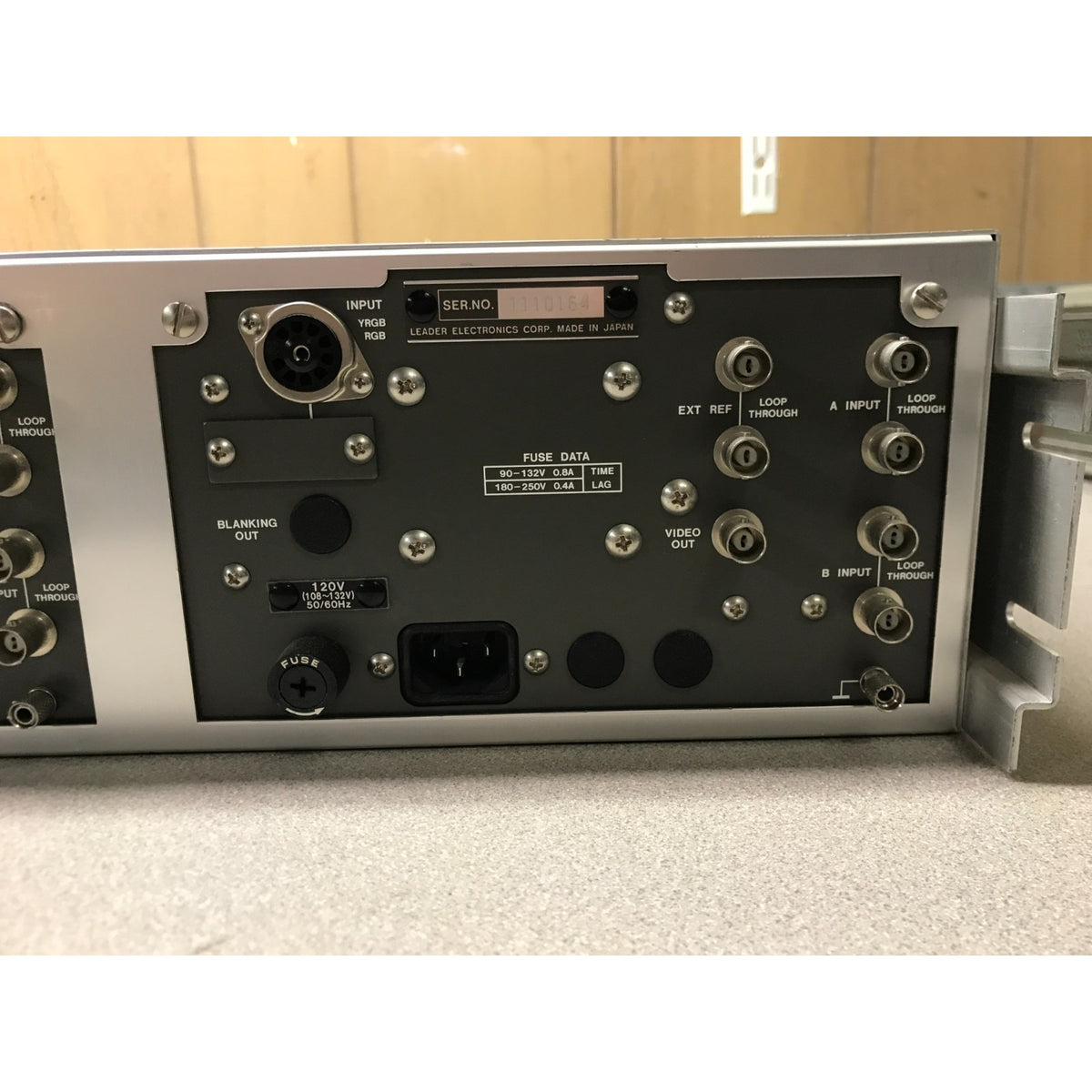 Close-up of the rear panel of a silver and black electronic signal generator or audio device, showing various input/output connectors, labels, and a power inlet on a tabletop.
