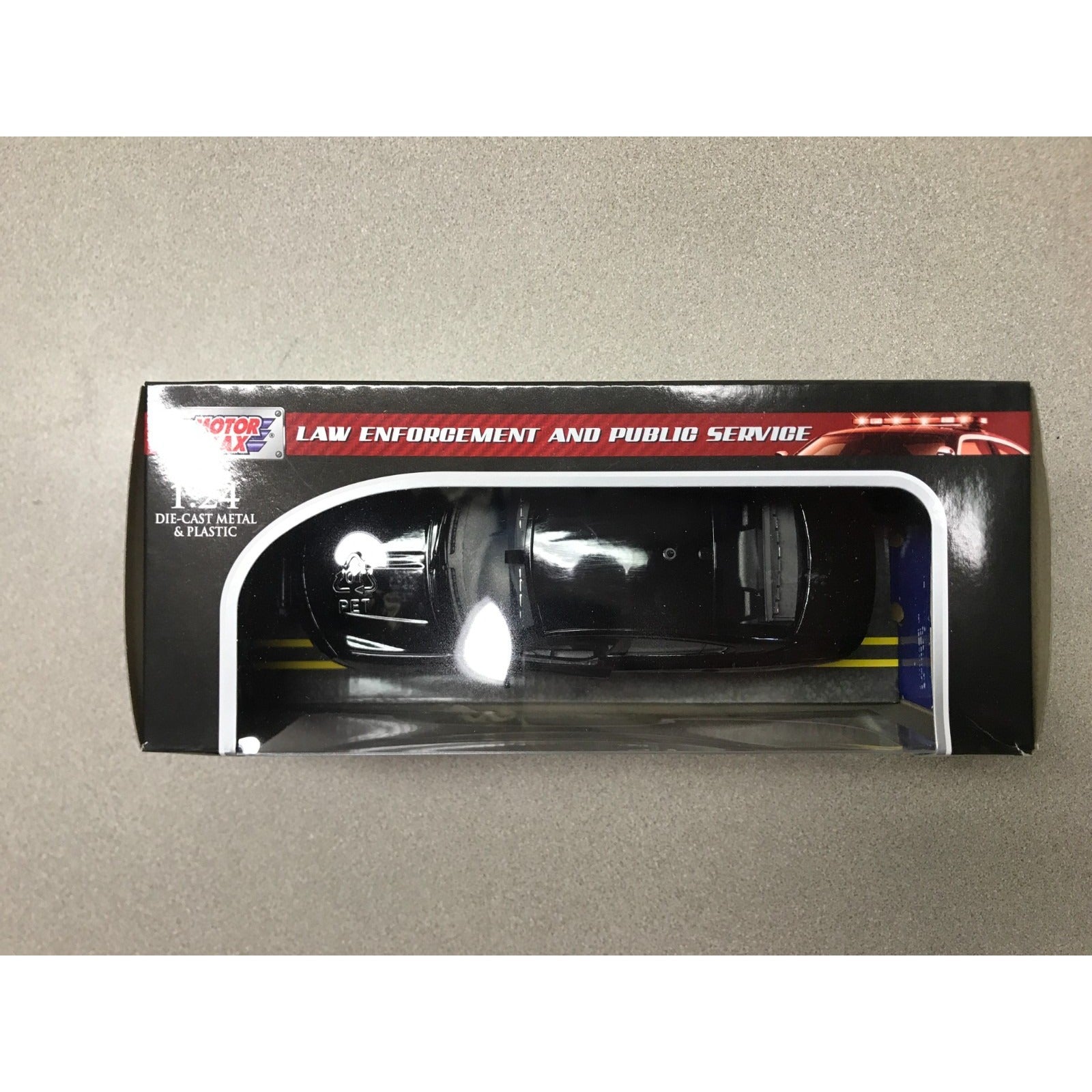A boxed die-cast metal model car in a black police-style vehicle, shown inside its retail packaging on a plain beige surface. The box has a clear window and visible branding at the top left. Top view of Custom NH State Police Unmarked - Motor Max 1:24 Die-Cast Car