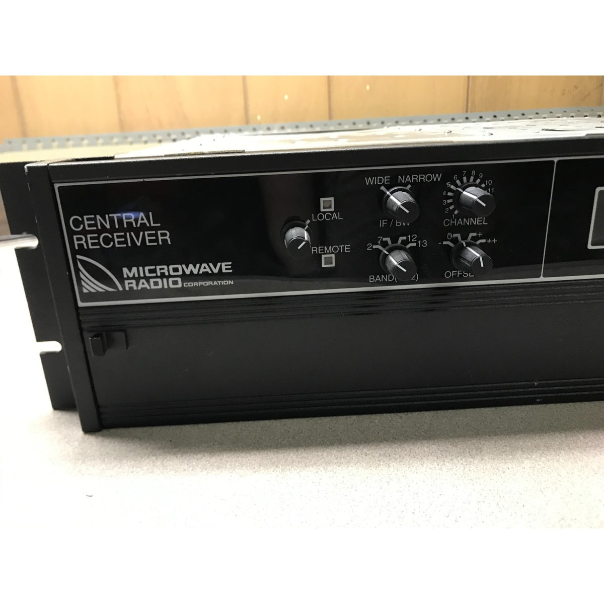 A black Microwave Radio Corporation Central Receiver radio amplifier control unit with multiple rotary knobs and white text labels, photographed on a light surface with a wood panel background.