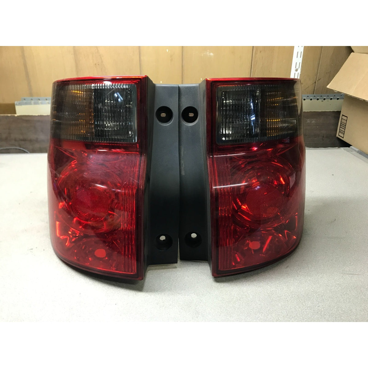 A pair of red tailgate light assemblies with black housings, photographed on a light gray surface with a cardboard box and wooden wall in the background. 2003-2008 Honda Element Tail Light Assembly - Left and Right