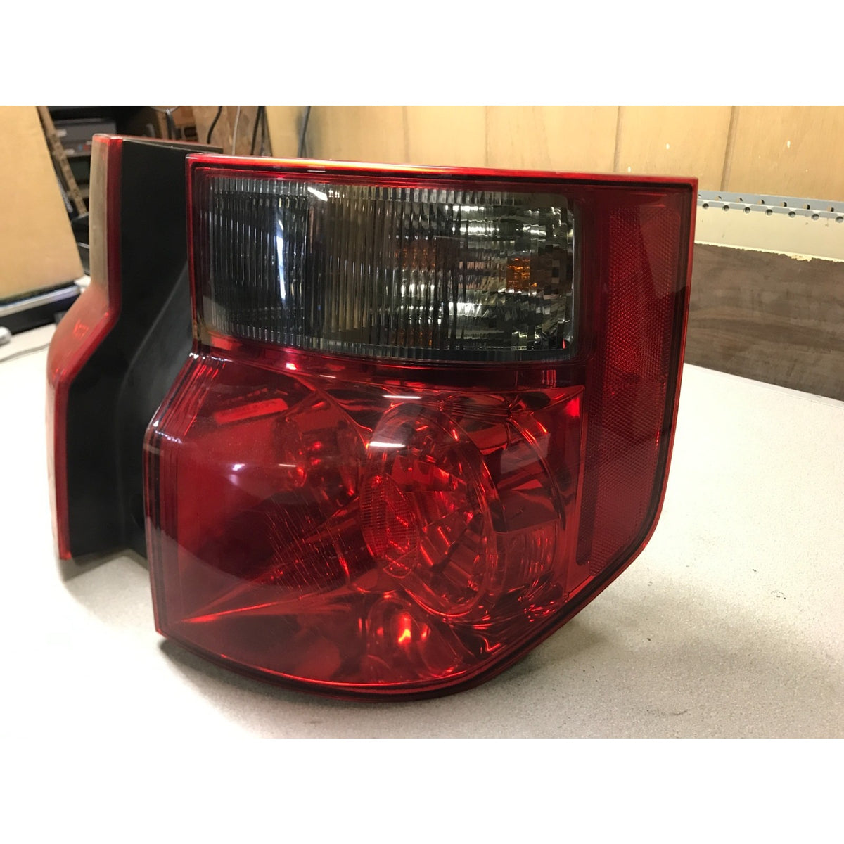 A red and black automotive tail light assembly photographed on a light-colored surface with a workshop or garage background. 2003-2008 Honda Element Tail Light Assembly  Right