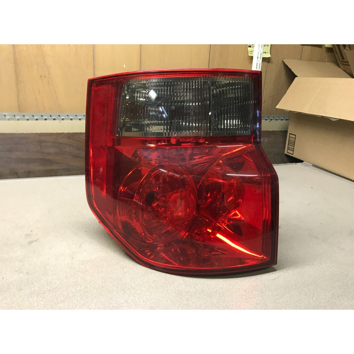 A red plastic vehicle tail light assembly photographed on a light surface with a cardboard box and wood wall in the background. 2003-2008 Honda Element Tail Light Assembly - Left