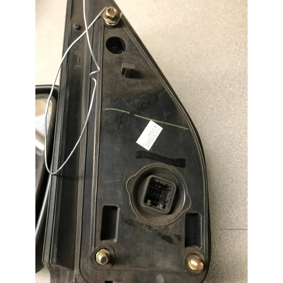 A used black plastic automotive side mirror housing with a connector port, mounting holes, and a wire, photographed on a light gray floor.