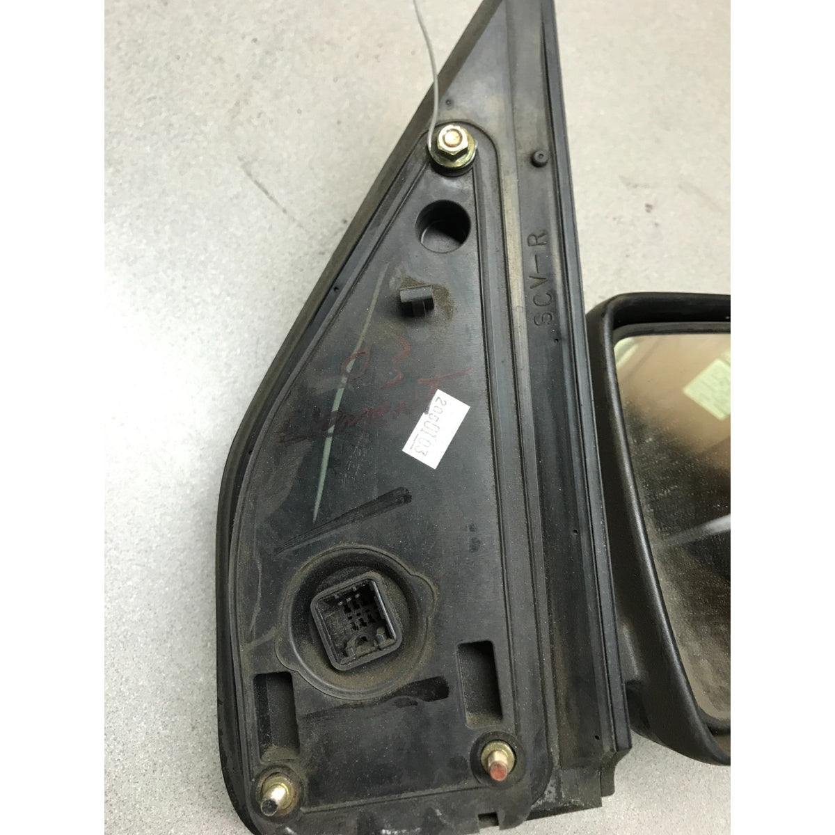 A used black plastic automotive side-view mirror housing with an attached mirror, photographed on a light gray concrete floor.