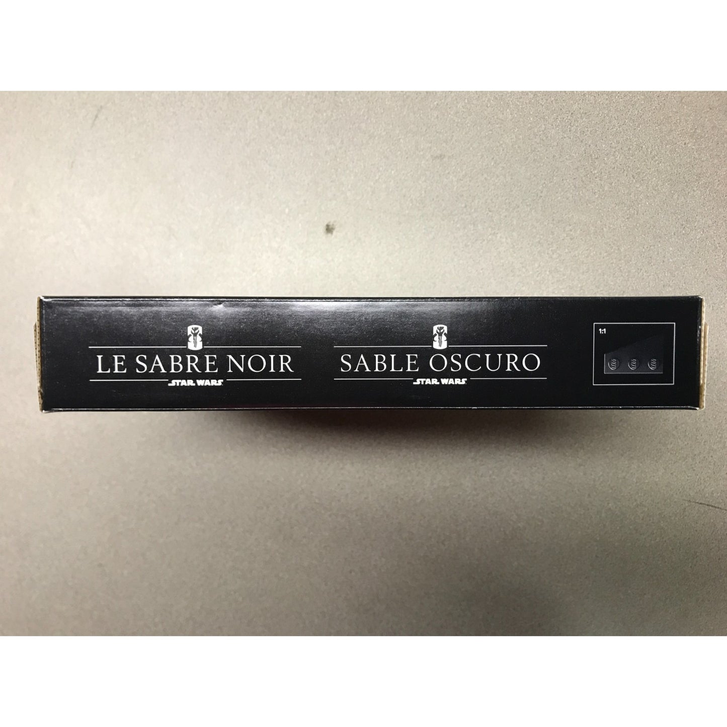 A long, narrow black product box with white French text reading 'LE SABRE NOIR' and 'SABLE OSCURO' plus 'STAR WARS' logos, photographed on a plain light gray surface.