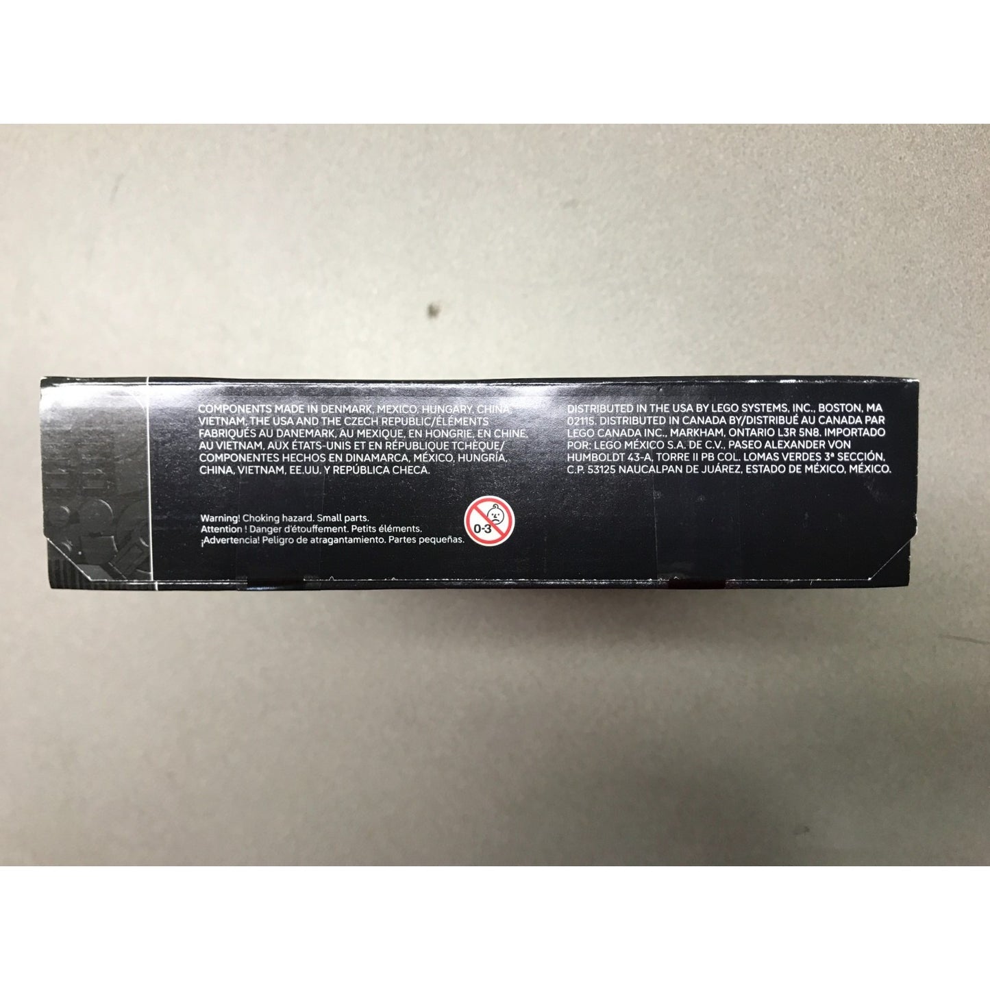 The side of a black LEGO box with white printed text and a circular warning symbol, photographed on a plain light gray surface.
