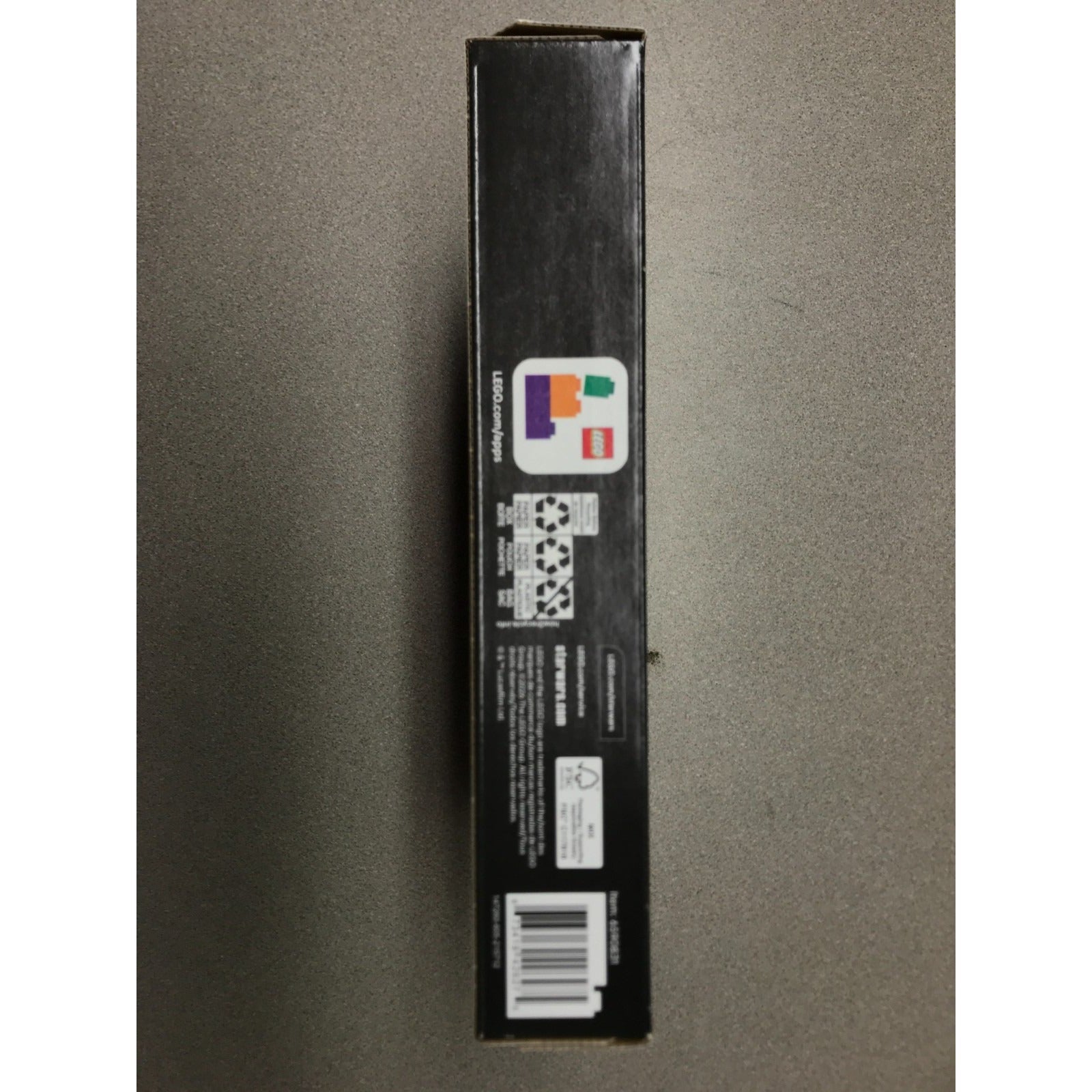 A tall, narrow black product box standing upright against a gray background, showing printed labels, a barcode, and a small multicolor logo near the top.