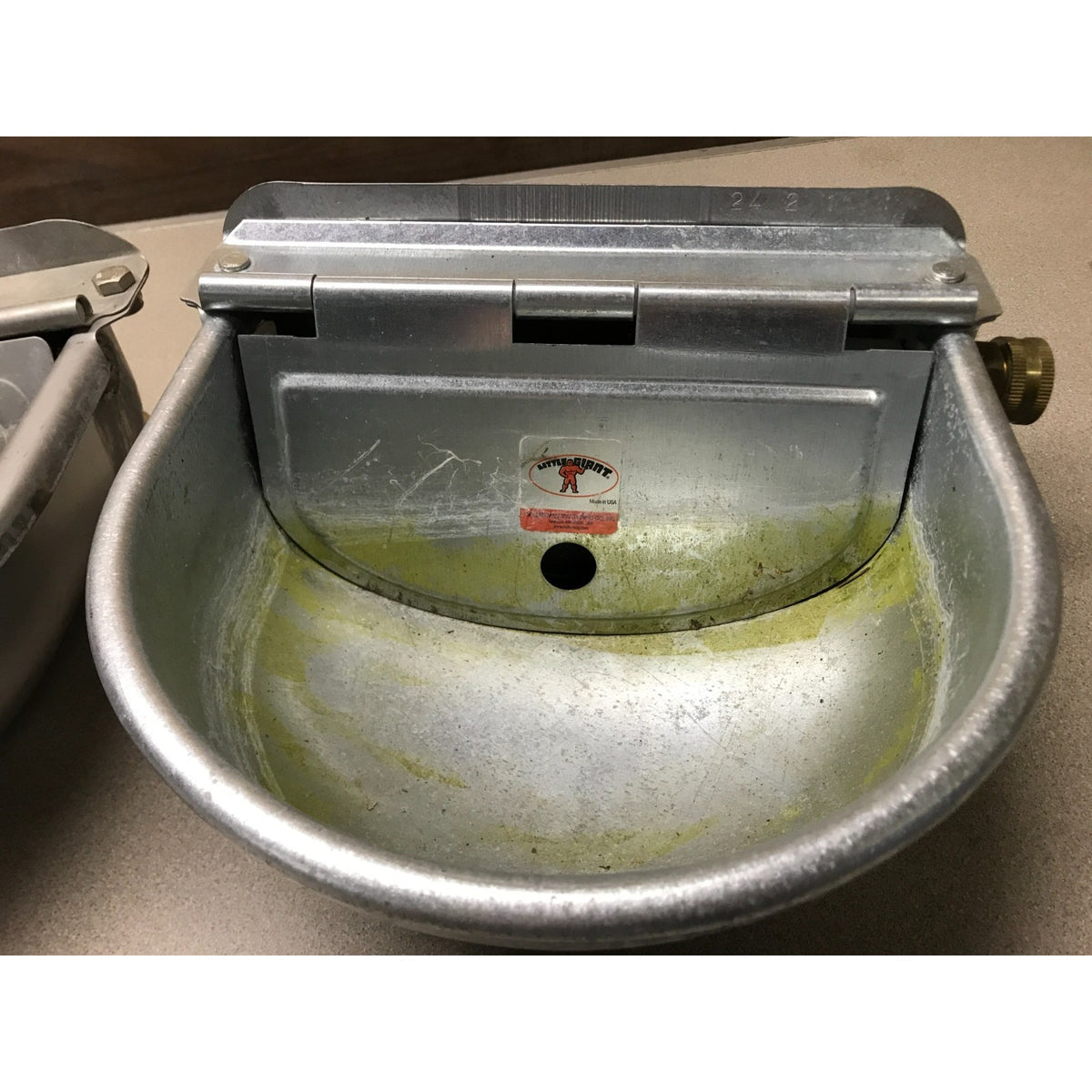 A stainless steel commercial sink or drain basin with a hinged lid, showing a yellow-green residue or buildup inside the bowl and a small red and white brand sticker in the center.