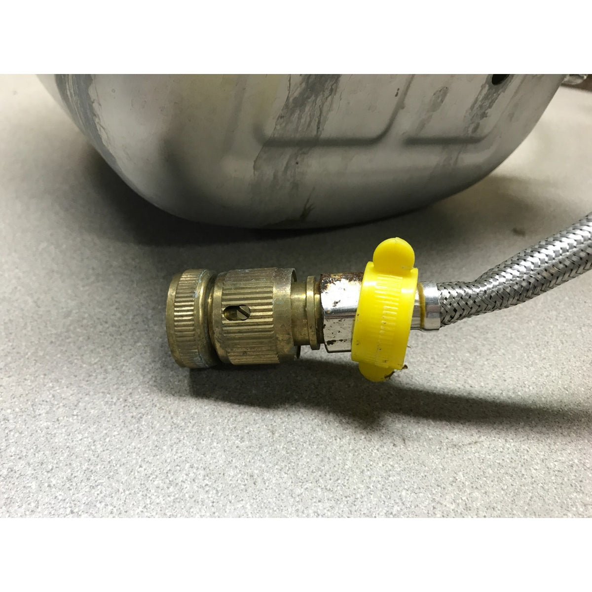 A close-up of a brass gas adapter with a yellow plastic cap and a braided metal hose, positioned near the base of a silver metal container on a gray surface.