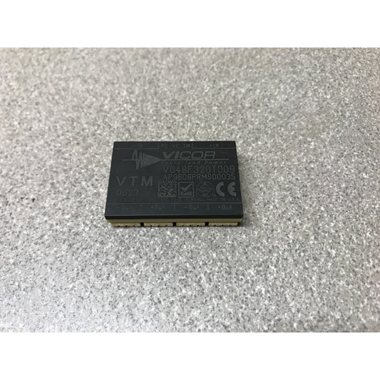 A Vicor VTM 24V DC 100W power module with gold-plated pins, resting on a textured gray surface.
