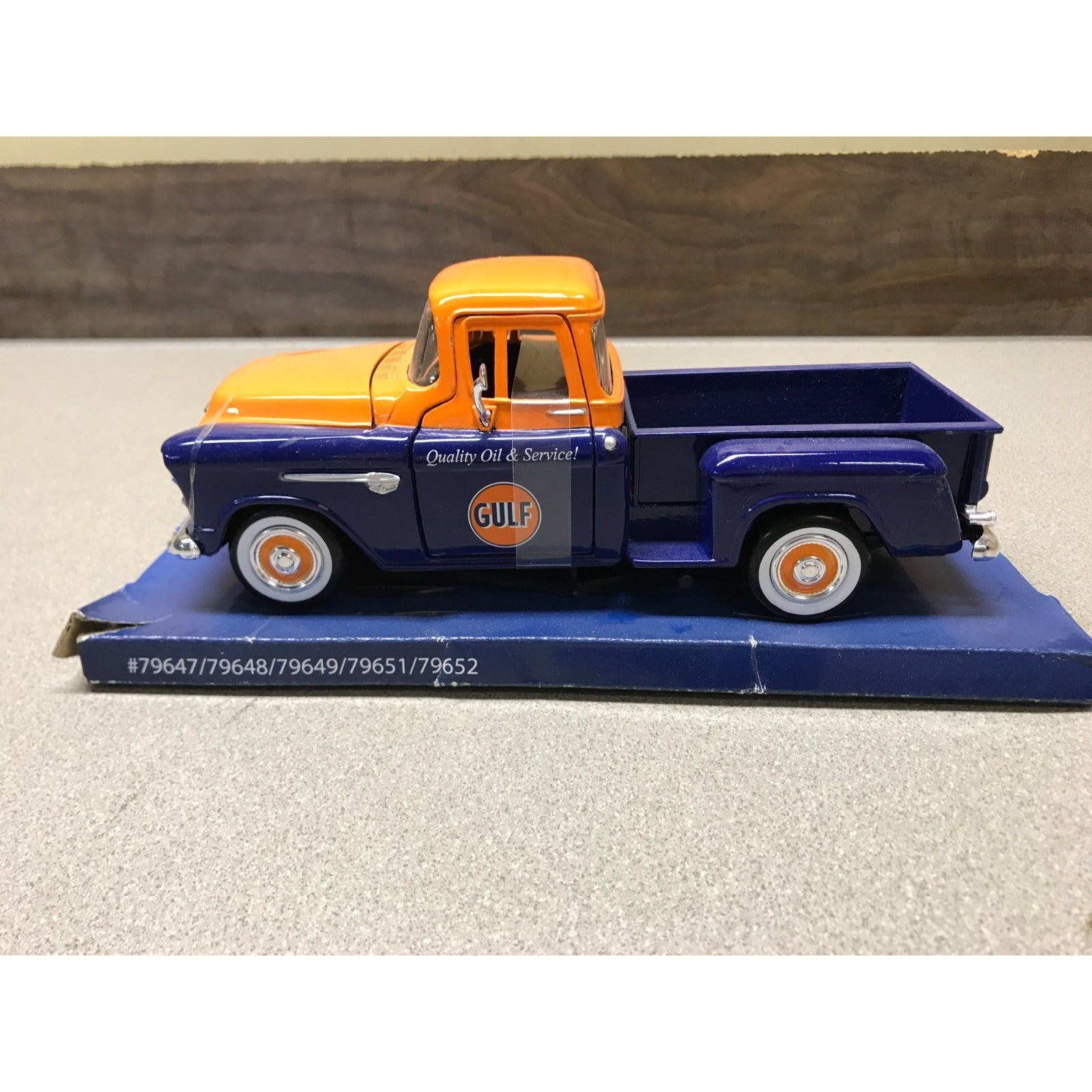 A vintage-style toy pickup truck displayed on a blue base. The truck has an orange cab and hood, a dark blue body, and orange-and-white wheels.