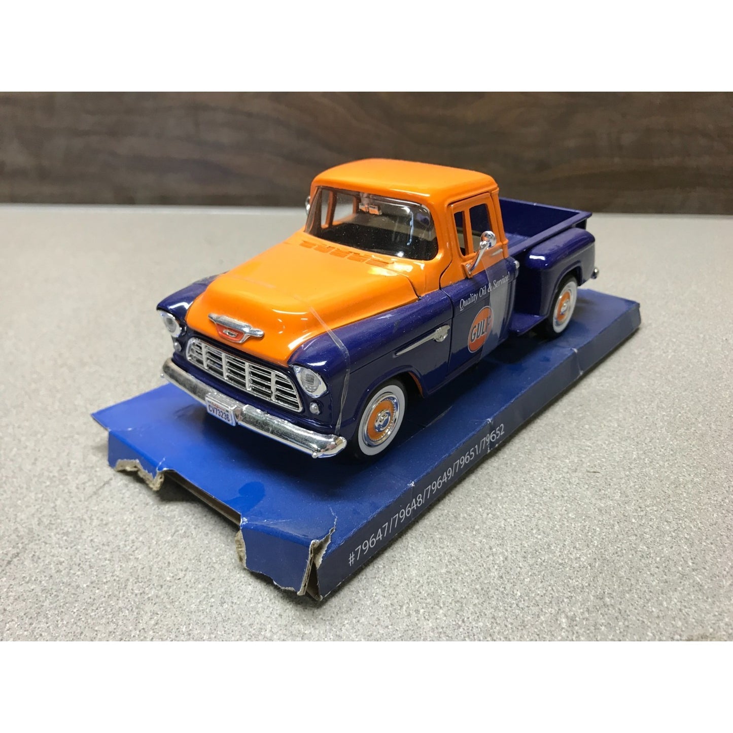A vintage-style toy pickup truck model painted orange and dark blue, displayed on a blue base against a neutral gray surface with a dark wood background.