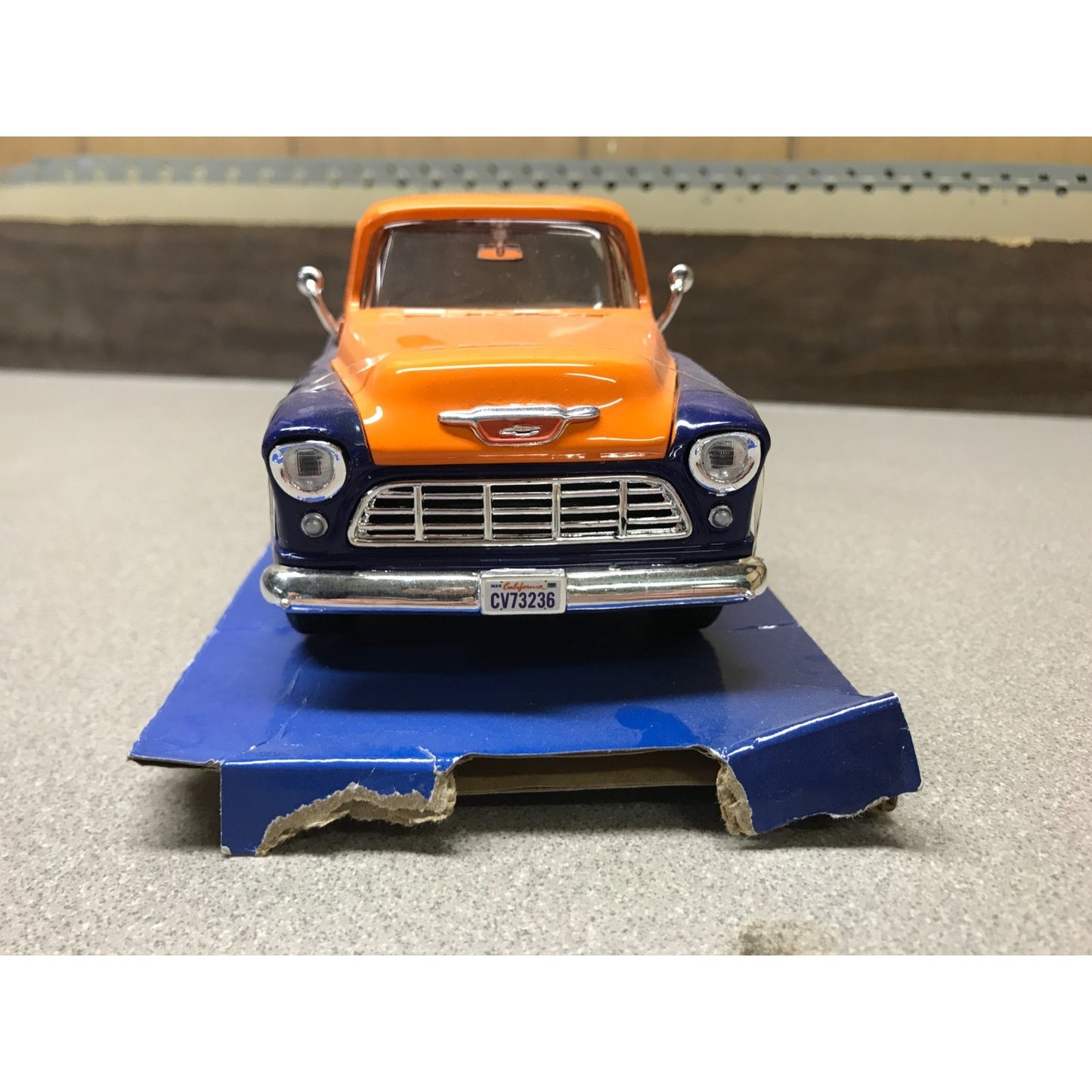 A vintage-style toy or model car in orange and dark blue, displayed on a blue base against a neutral indoor background.