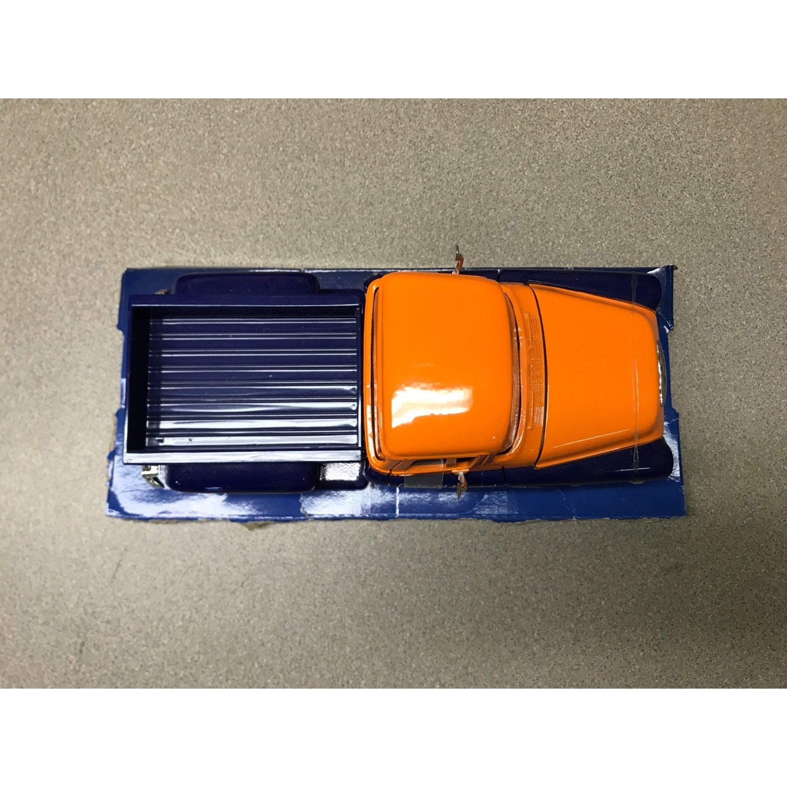 A miniature orange and blue toy pickup truck, viewed from directly above, resting on a textured gray surface.
