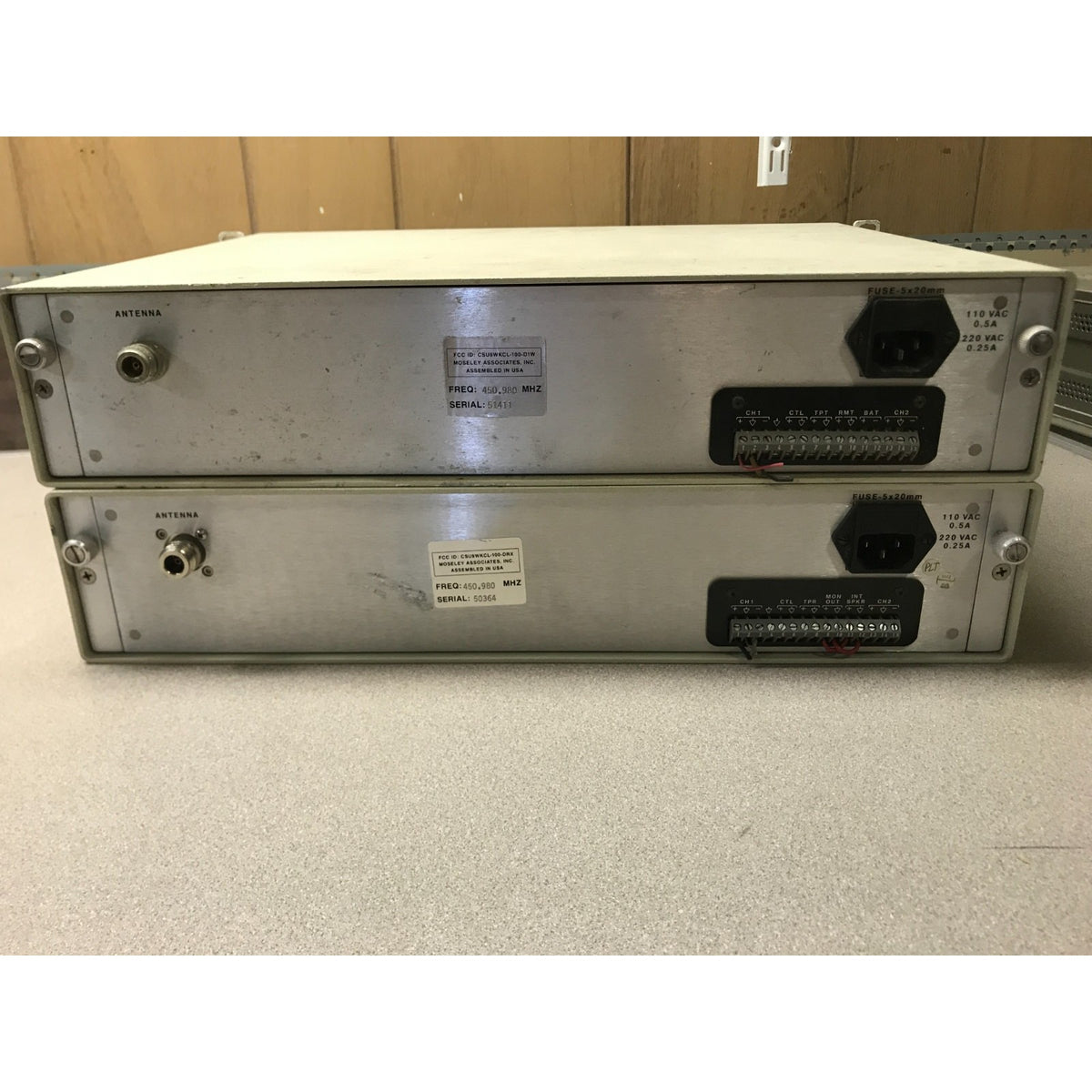 Two stacked industrial electronic modules with silver metal casings and black terminal blocks, photographed on a light floor against a wood-paneled wall. Moseley CL-100 Communications Link back panel view