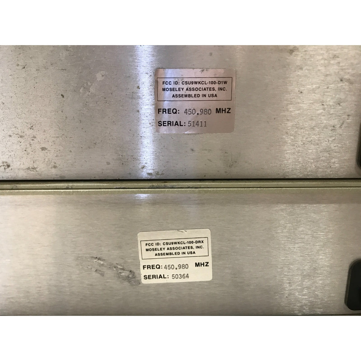 Close-up of a brushed metal appliance or equipment panel with two white product stickers showing FCC IDs, frequencies, and serial numbers. Moseley CL-100 Communications Link back panel