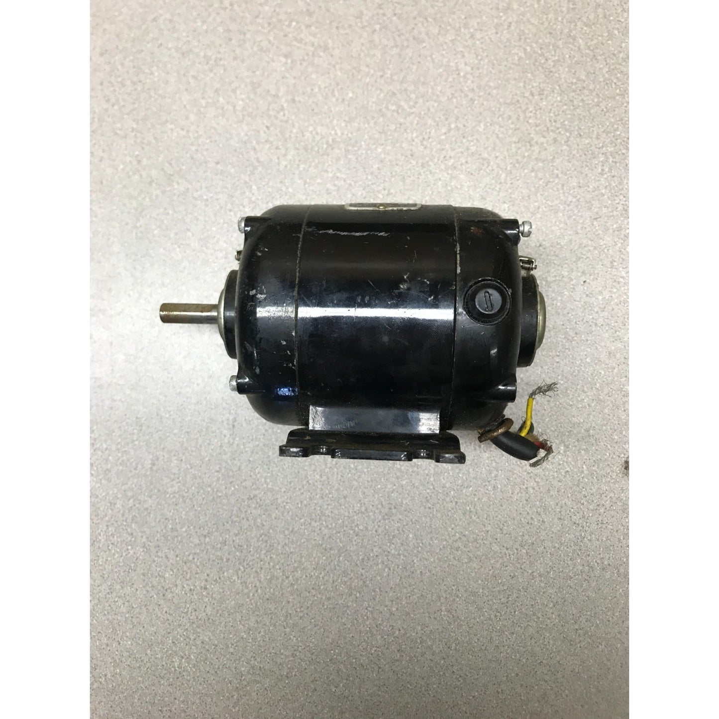 A black industrial electric motor with a metal shaft and attached wiring, photographed on a textured light gray surface.