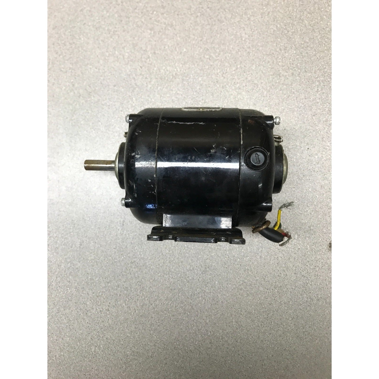 A black industrial electric motor with a metal shaft and attached wiring, photographed on a textured light gray surface.