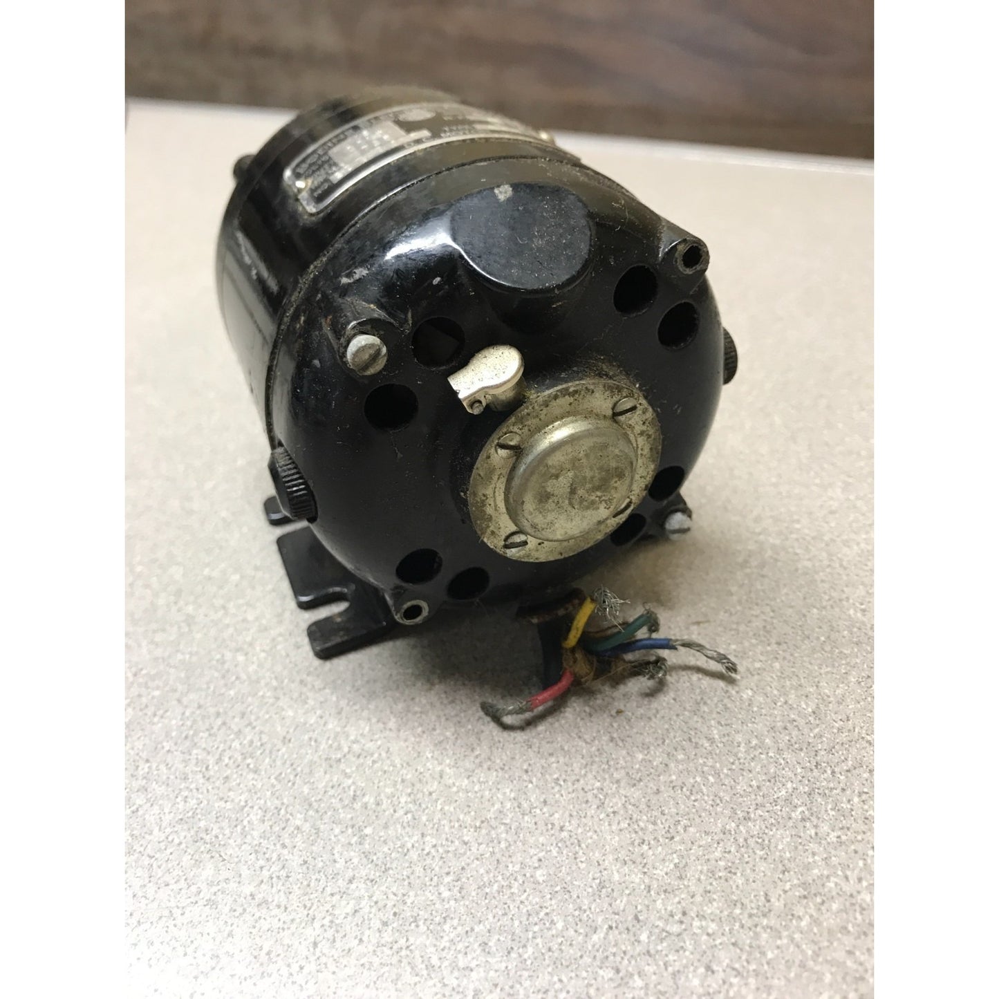 A used black electric motor with attached wiring, photographed on a light gray textured surface.