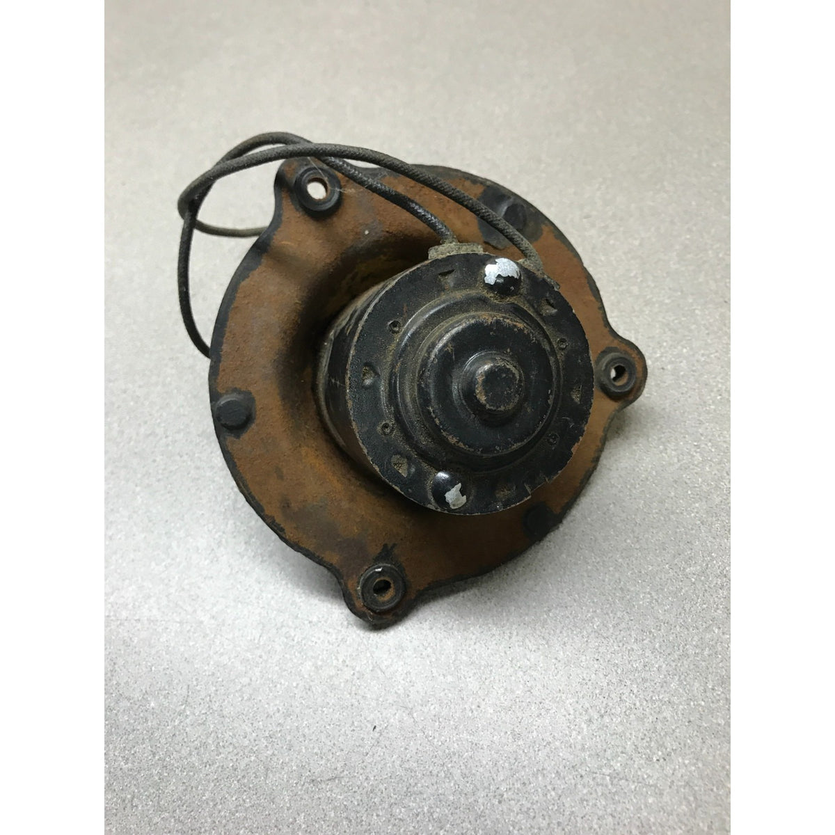 A used automotive or machinery component, likely a distributor, alternator, or starter motor part, featuring a rusted brown metal housing, black central assembly, and attached black wire leads, photographed on a neutral gray background.