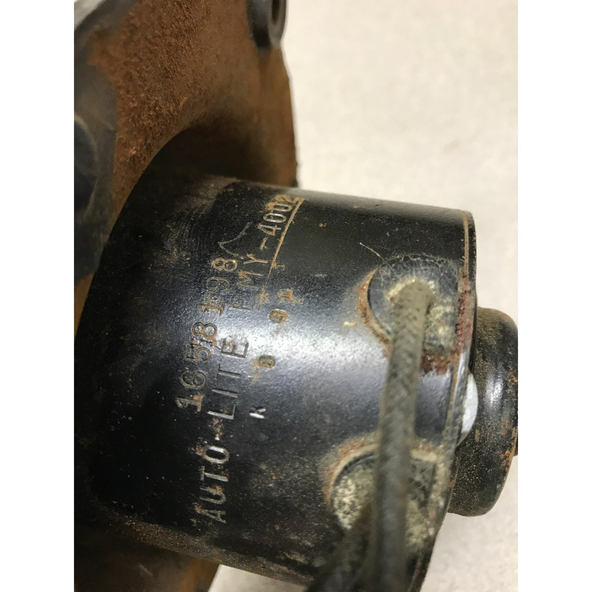 A close-up of a black cylindrical automotive part with stamped text reading 'AUTO-LITE' and '1076-638', showing rusted metal edges and a dark cord or strap attached.