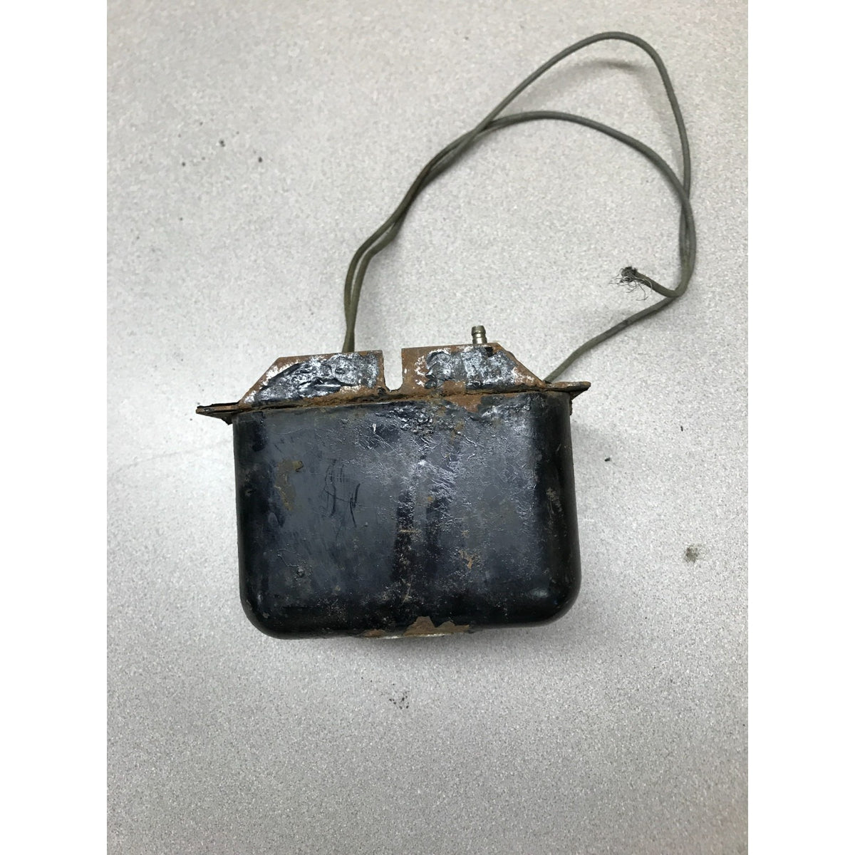 A used black rectangular automotive battery with a long wire lead, showing significant rust and wear on the top plates and casing, placed on a light gray textured surface.