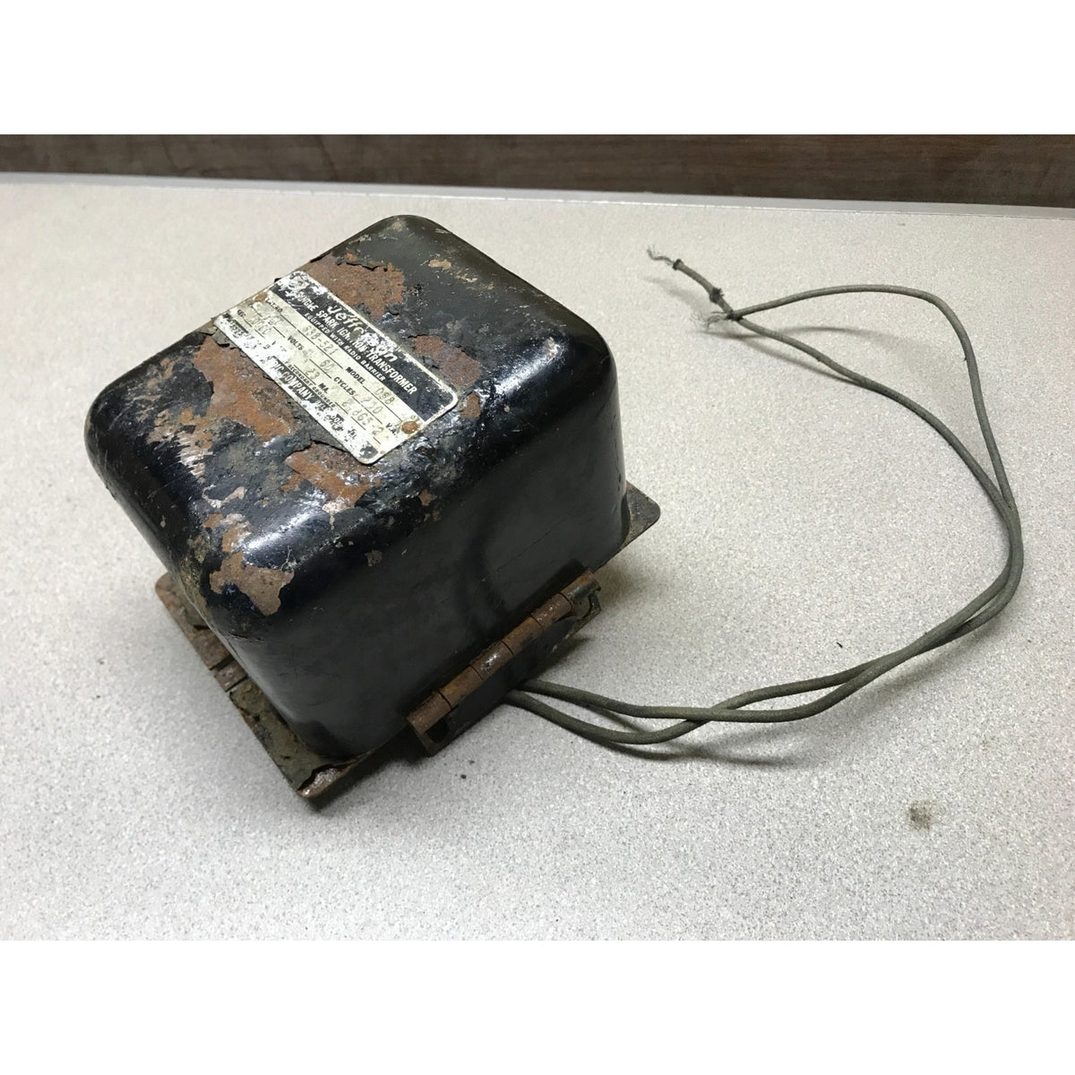 A used black metal electrical transformer with a weathered label and attached wires, resting on a light gray surface. Jefferson Single Spark Ignition Transformer Cat No. 638-521 Model No 058 Equipped with Radio Barrier
side view
