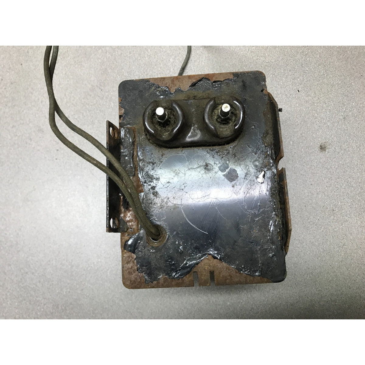 A used, weathered metal electronic component or transformer with two circular terminals on top and attached wires, photographed on a light gray surface.