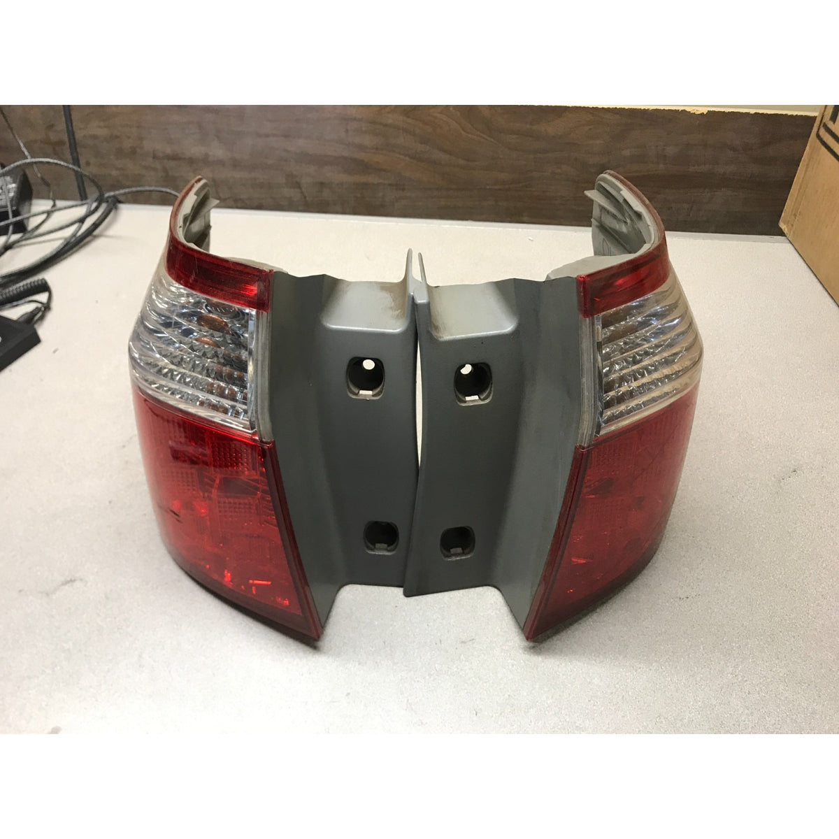 A pair of rear vehicle tailgate light assemblies with red lenses and silver/white reflectors, photographed on a light gray workbench surface with a wood panel background. Left and Right Tail Light Assemblies for 2005-2007 Honda Odyssey