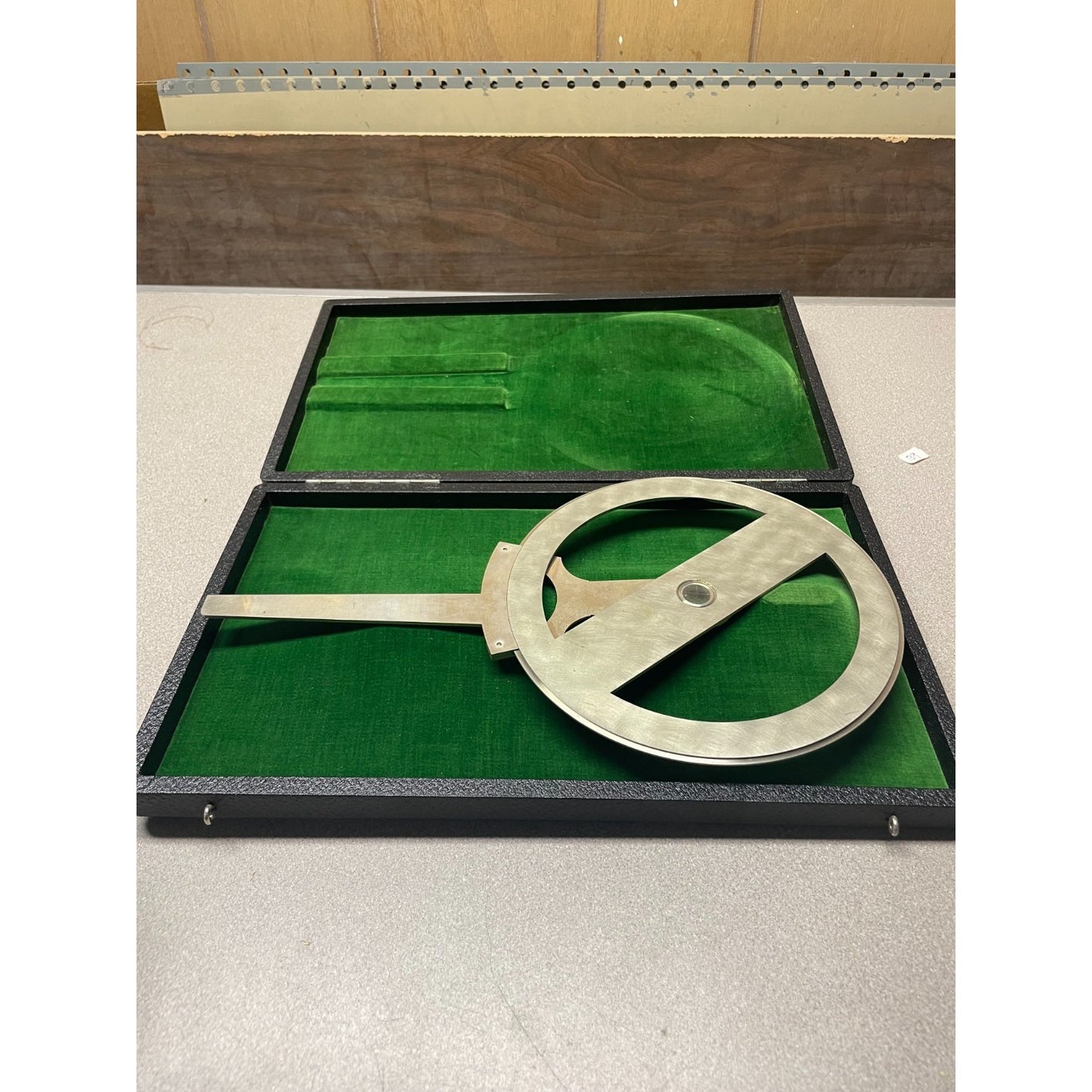 A vintage metal surveyor's square or angle measuring tool resting inside an open black presentation case lined with green velvet, set on a light gray tabletop.