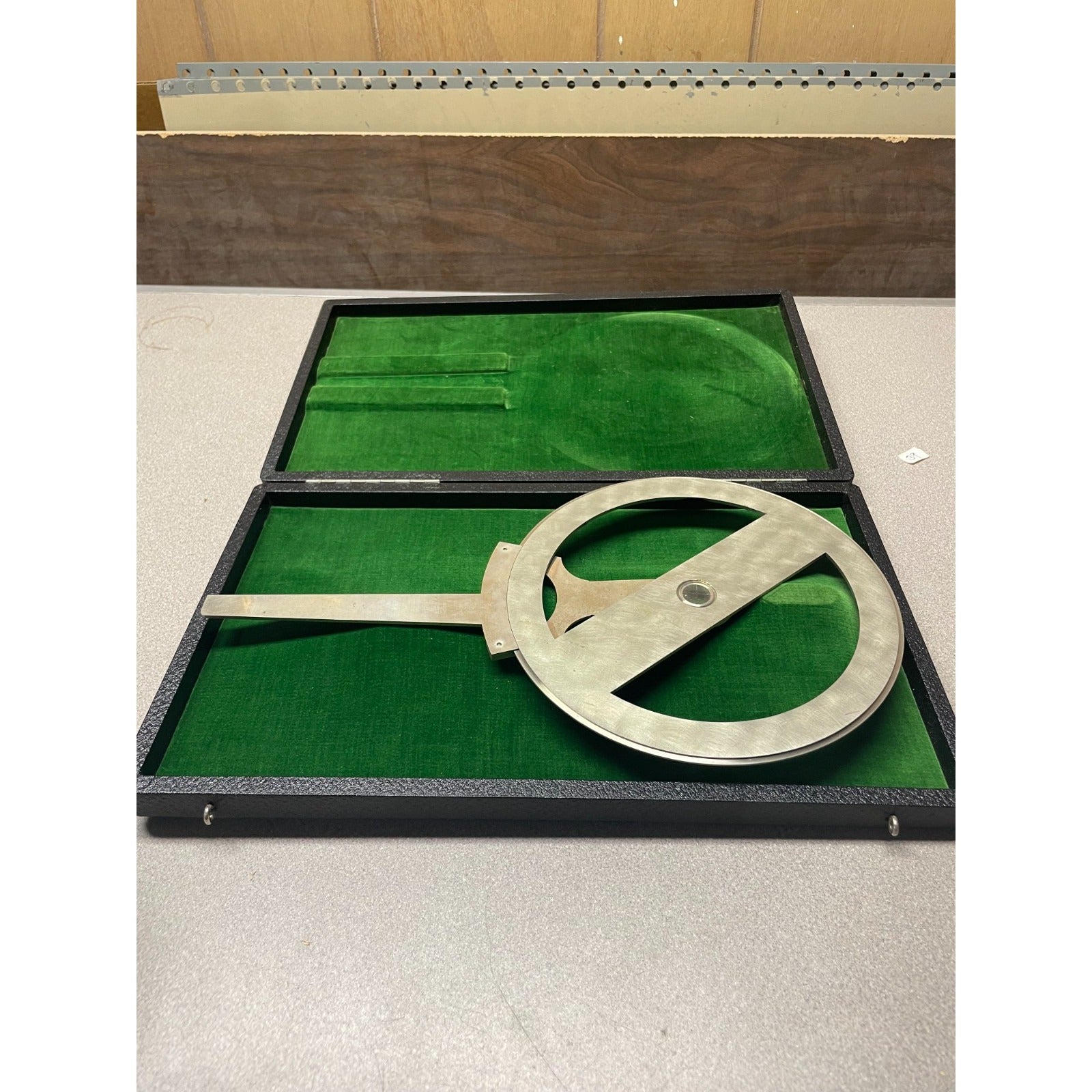 A vintage metal surveyor's square or angle measuring tool resting inside an open black presentation case lined with green velvet, set on a light gray tabletop.
