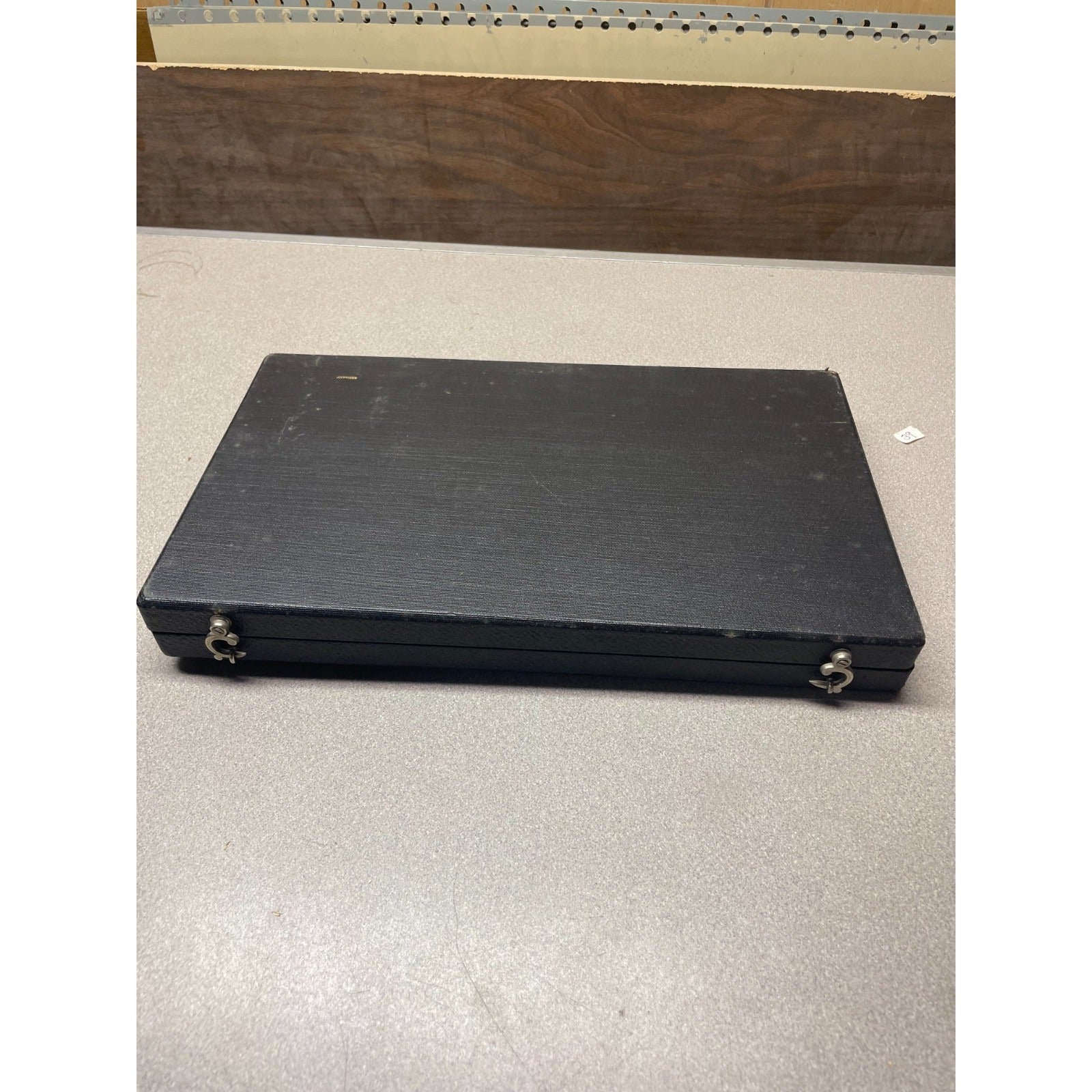 A rectangular black hard case with two small metal ring handles, photographed on a light gray floor in front of a wooden panel.